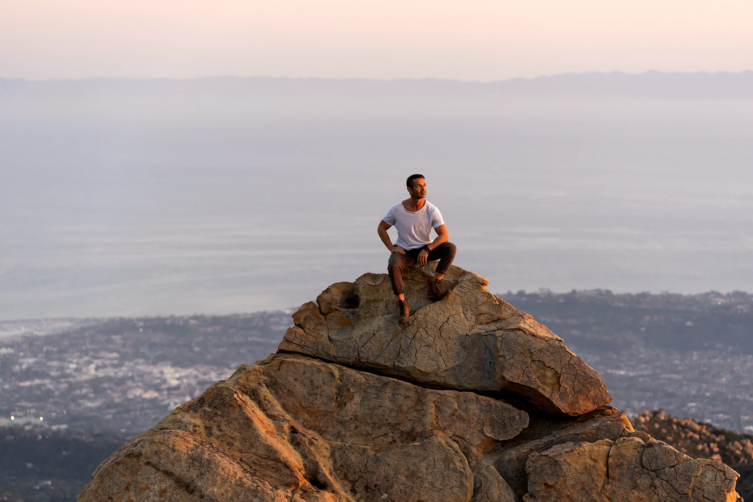 John Burgos sitting on a sandstone summit at sunset, the coastline and ocean stretching out below him