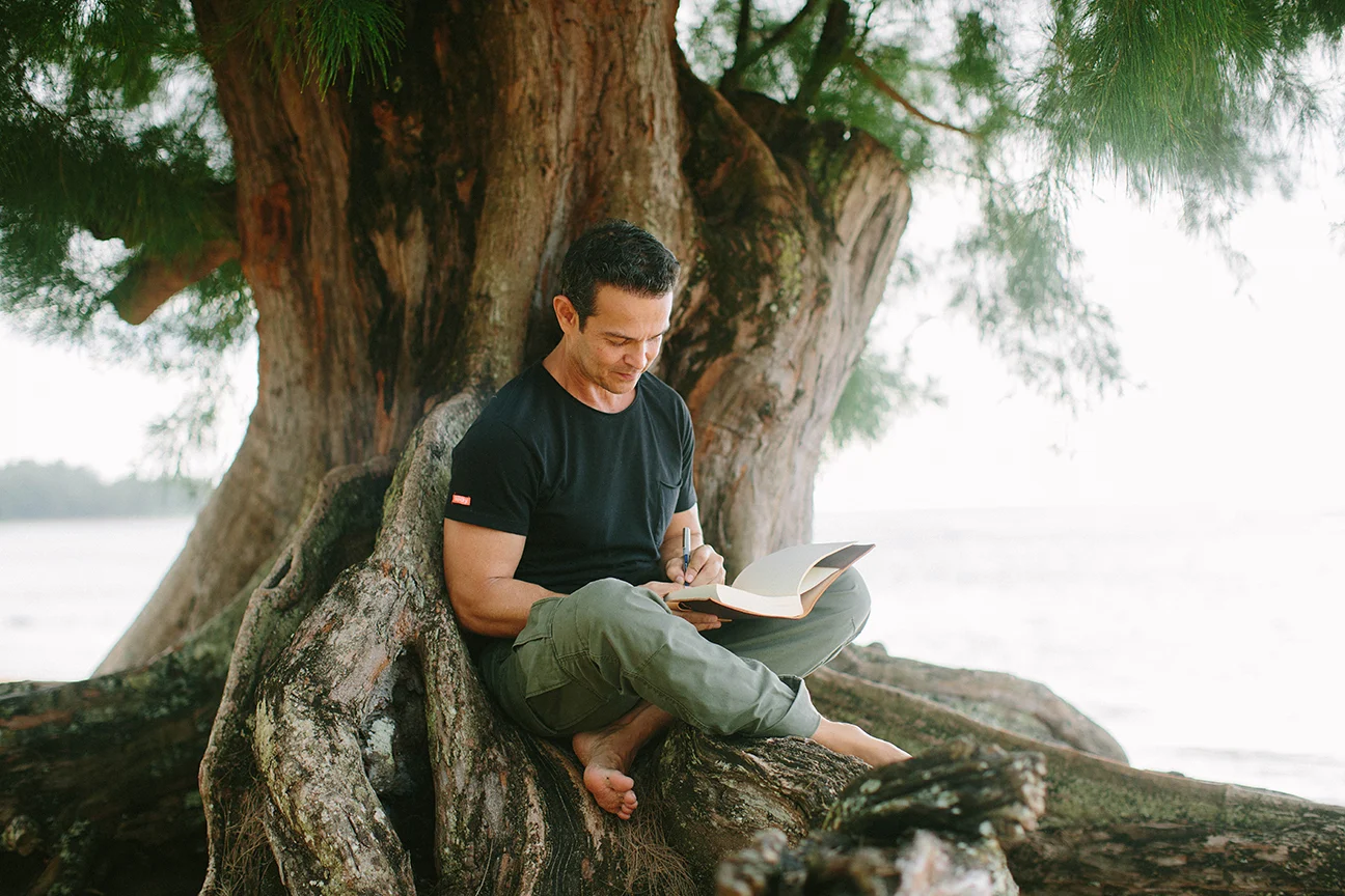 John Burgos writing in his journal, seated barefoot among the roots of a large tree by the ocean