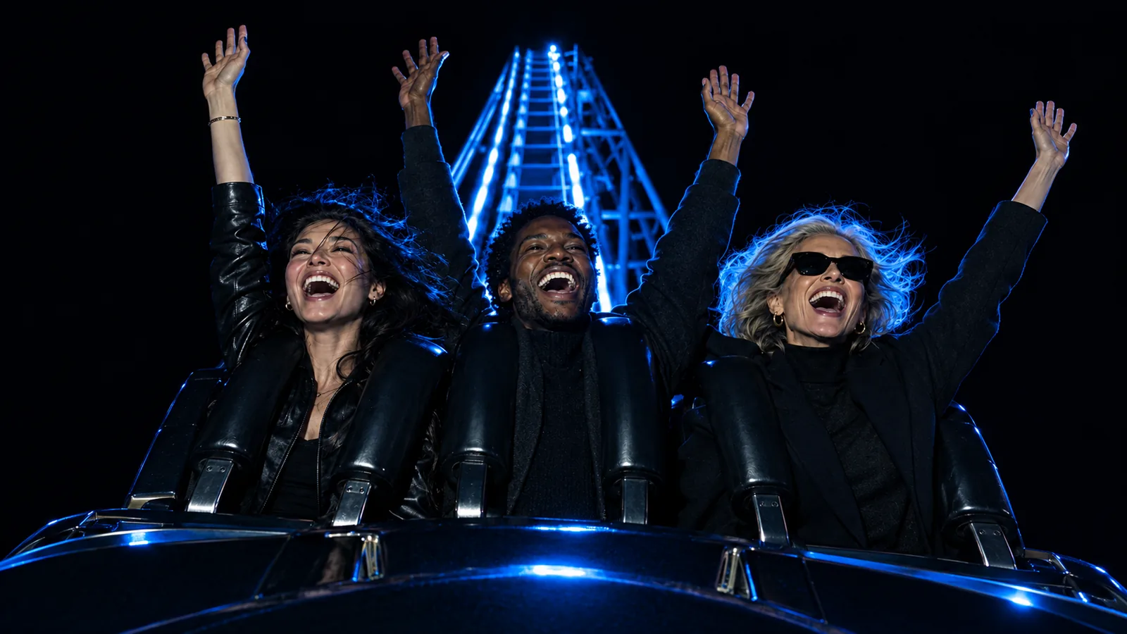 Three friends with arms raised, laughing at the crest of a roller coaster at night under blue light